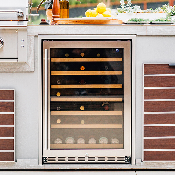 A built-in outdoor wine refrigerator with a glass door and stainless steel trim is integrated into a light grey stone kitchen island. The cooler features wooden shelving holding wine bottles and a digital temperature display showing 55 degrees, flanked by dark wood-slat cabinet doors and a partial view of a stainless steel grill.